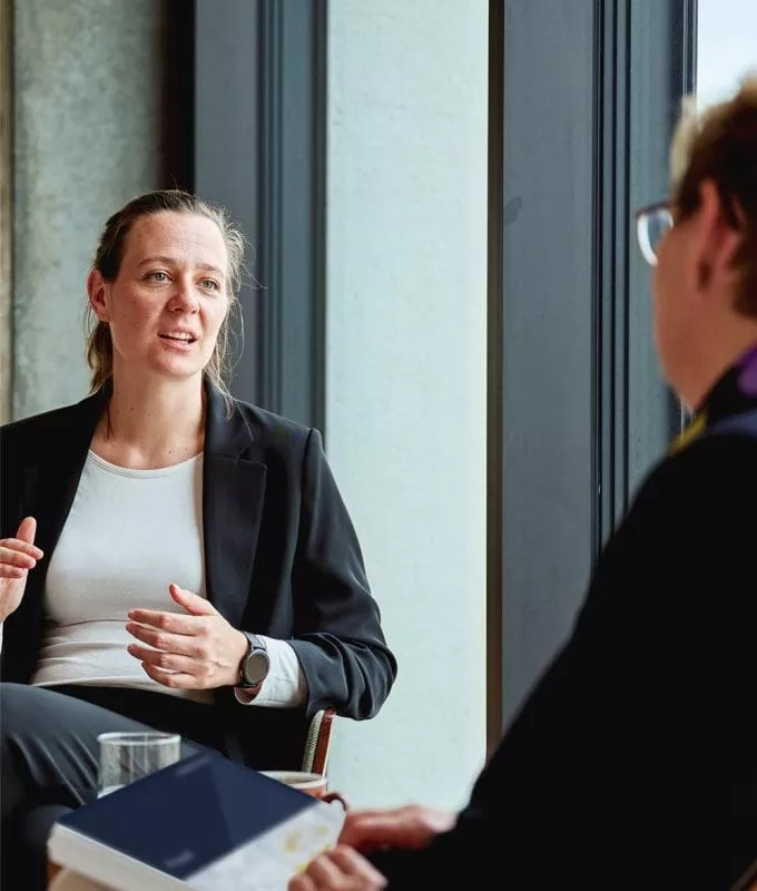 Two professionals having a conversation in a modern office setting, with one woman in a black blazer speaking and the other person listening attentively.