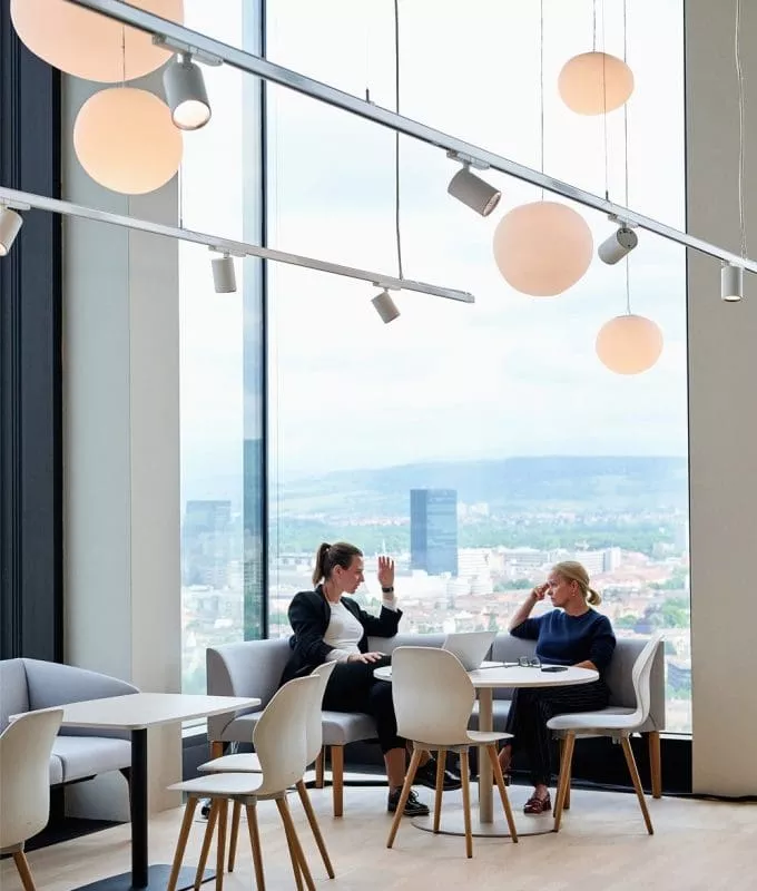 Two women seated at a round table having a discussion in a modern office lounge with large windows overlooking a cityscape.