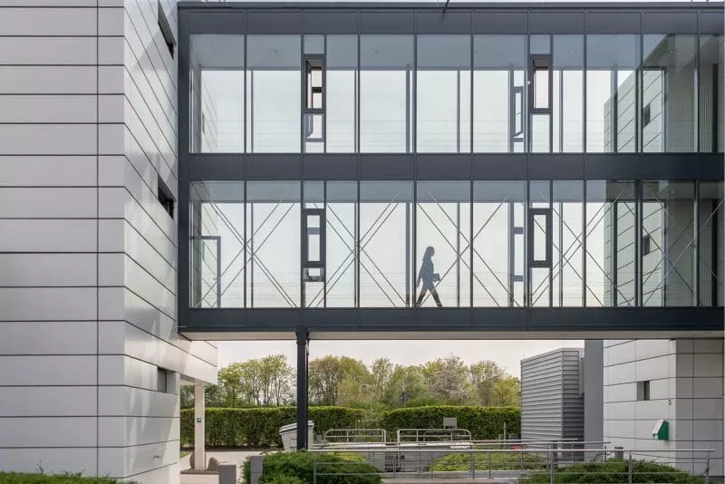 Person walking through a modern glass skywalk connecting two office buildings, with greenery visible in the background.