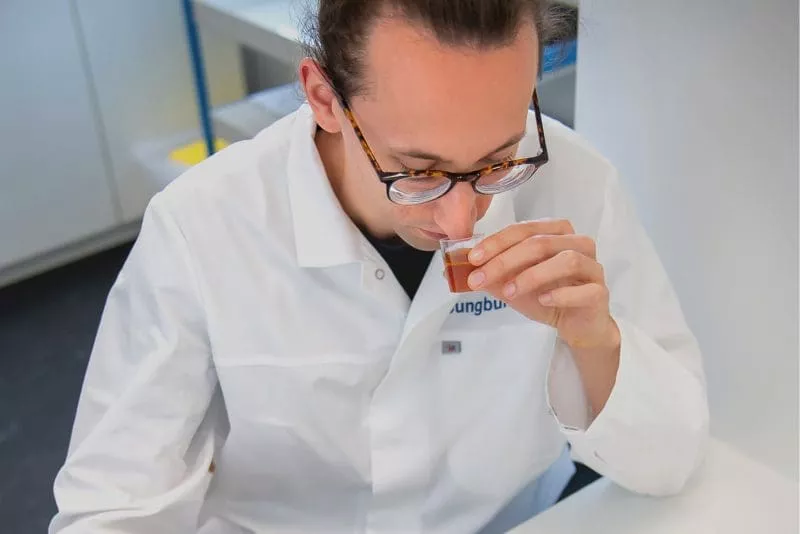 Scientist in a white lab coat smelling a small cup of brown liquid