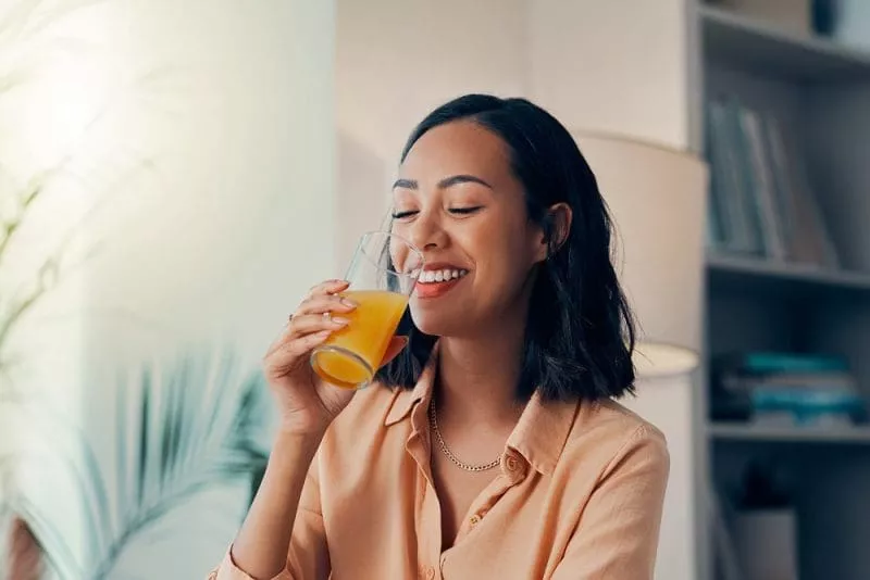 Smiling woman in a peach shirt drinking a glass of orange juice indoors.