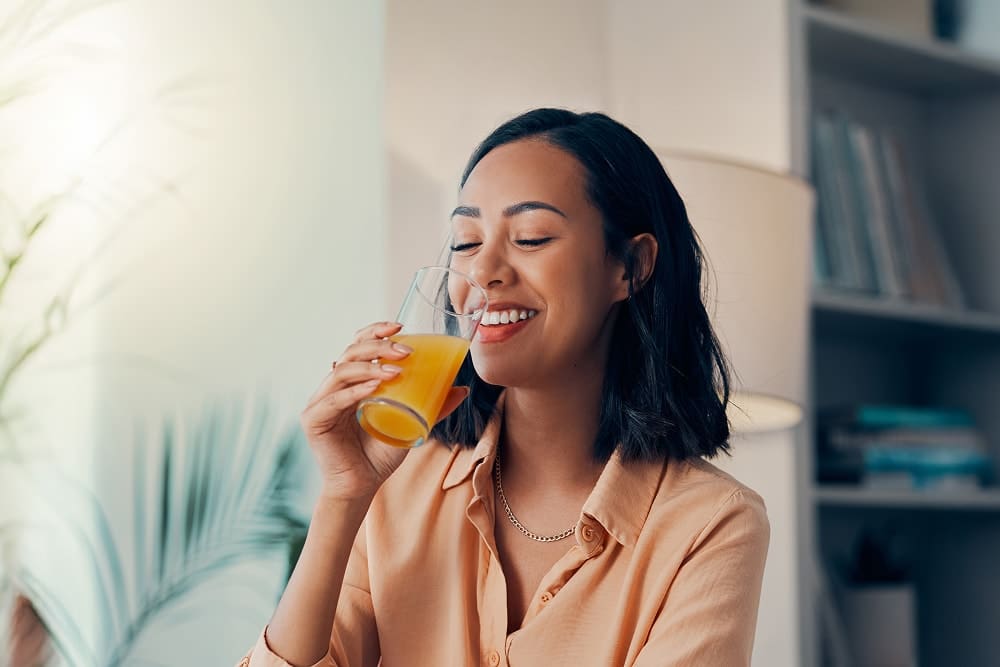 Smiling woman in a peach shirt drinking a glass of orange juice indoors.