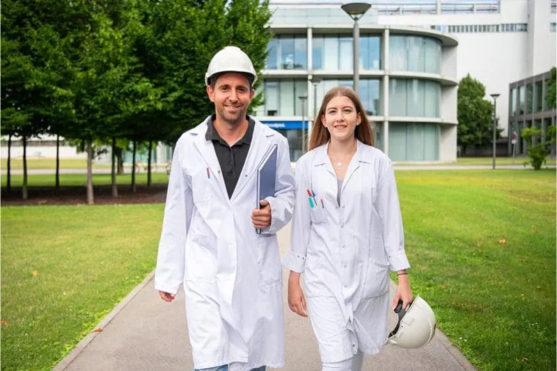 Two scientists in white lab coats walking outdoors on a path through a green campus, one holding a helmet and the other wearing one.