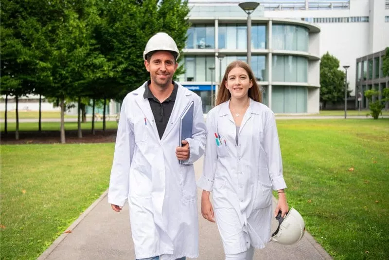 Two scientists in white lab coats walking outdoors on a path through a green campus, one holding a helmet and the other wearing one.