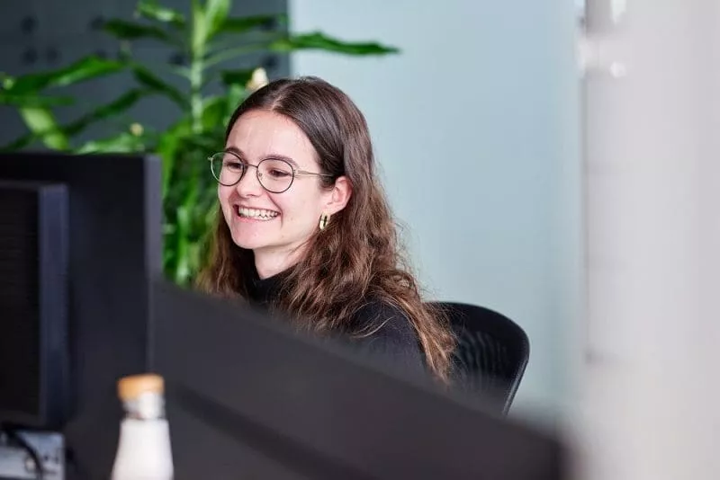 Smiling woman with long brown hair and glasses working at a computer in a bright office, with a green plant in the background.