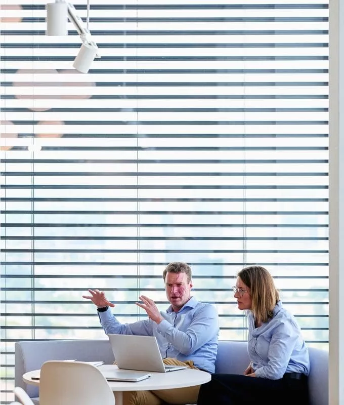 Man and woman in business attire sitting at a round table with a laptop, having a discussion in a modern office space with large windows and horizontal blinds.