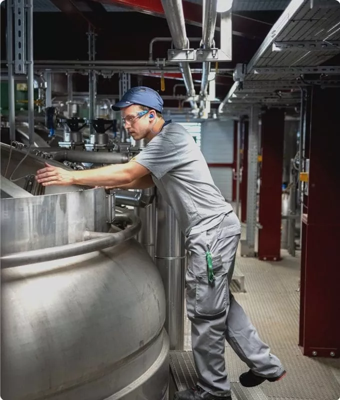 Industrial worker wearing safety gear inspecting a large stainless steel tank in a factory setting with complex piping and metal structures.