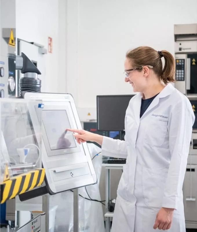 Female scientist in a lab coat using a touchscreen interface in a laboratory setting, smiling while interacting with the equipment.
