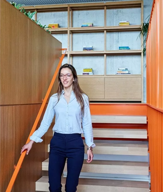 Woman walking down a modern indoor staircase with orange railings, in a bright office space with wooden bookshelves and contemporary design.