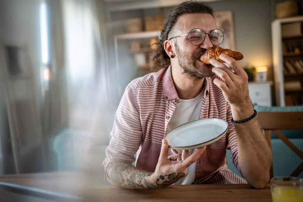 Smiling man with glasses and tattoos eating a croissant while holding a plate. image