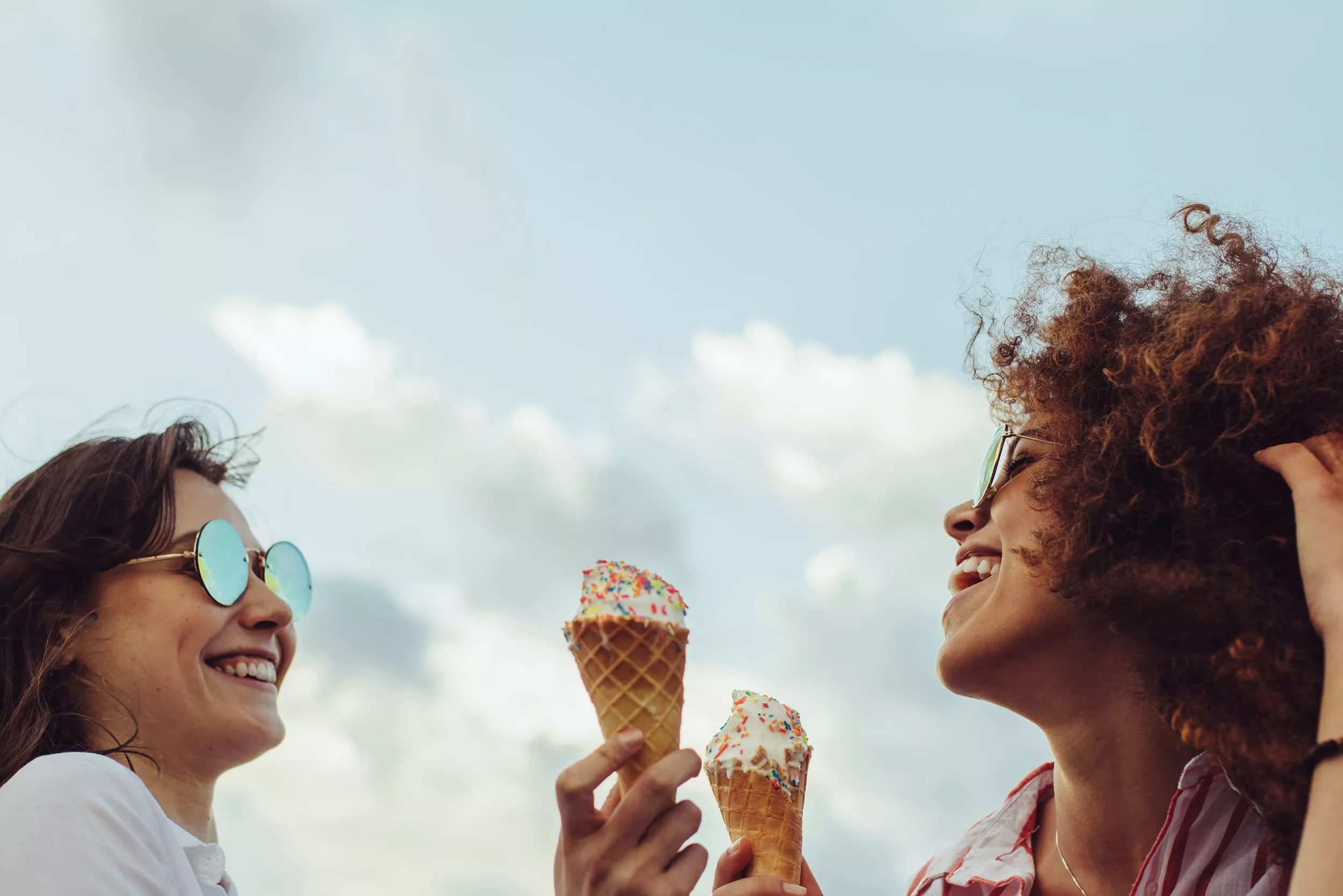 Two women smiling and holding ice cream cones with sprinkles against a cloudy sky. image