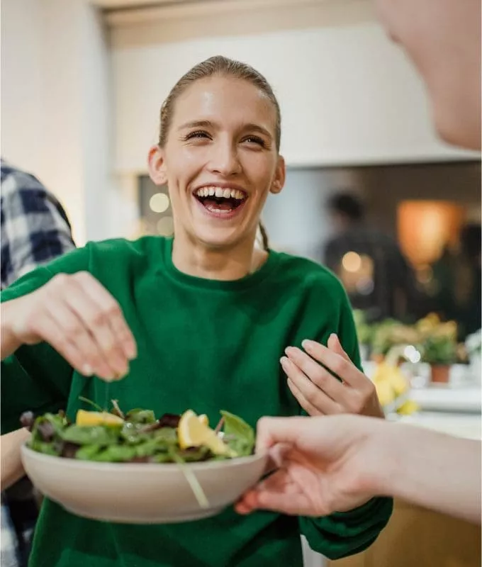 Smiling woman in green sweater being handed a salad bowl in a kitchen.
