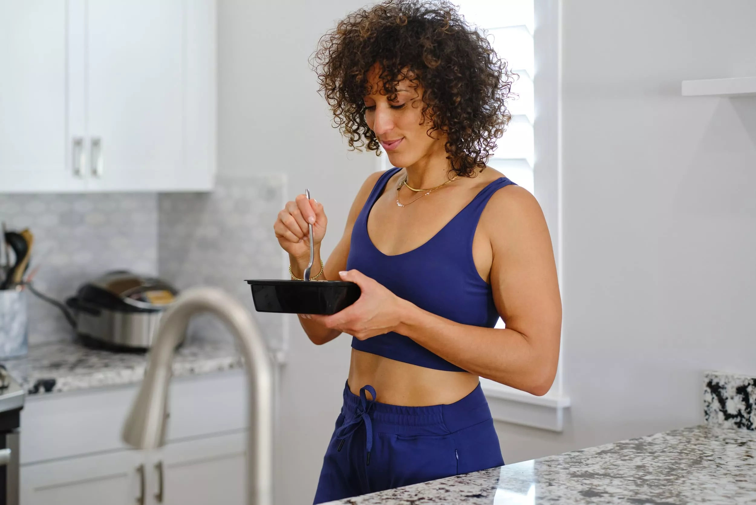 Woman in workout clothes eating from a black container in a modern kitchen.