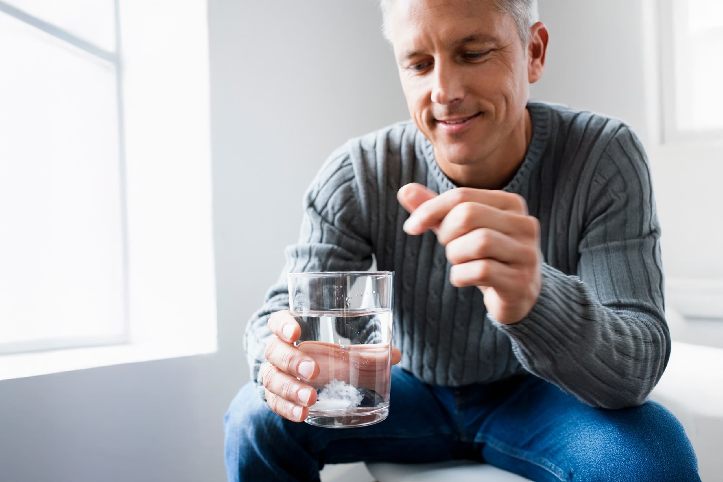 Man in grey sweater watching a tablet dissolve in a glass of water.