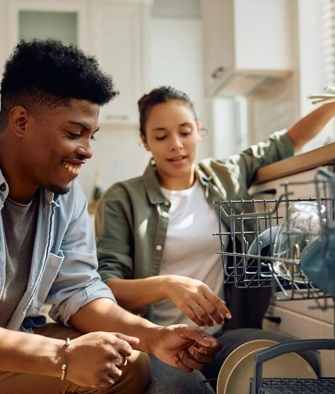 Smiling couple unloading clean dishes from a dishwasher in a sunlit kitchen.
