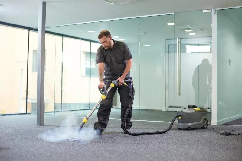 Man using a steam cleaner on carpet in a modern office with glass walls.