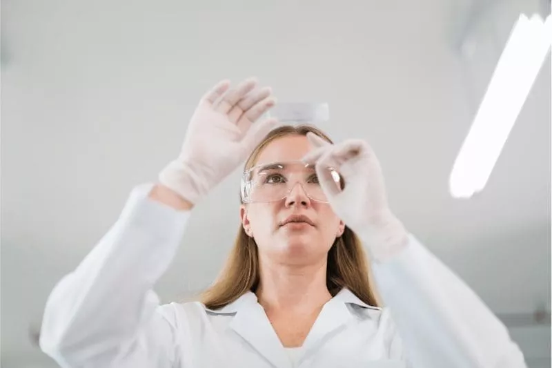 Female scientist wearing safety glasses and gloves inspecting a microscope slide in a laboratory.