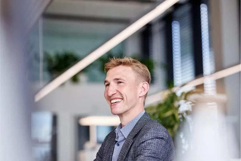 Smiling man in a checkered blazer and blue shirt in a bright, modern office with plants and large windows in the background.