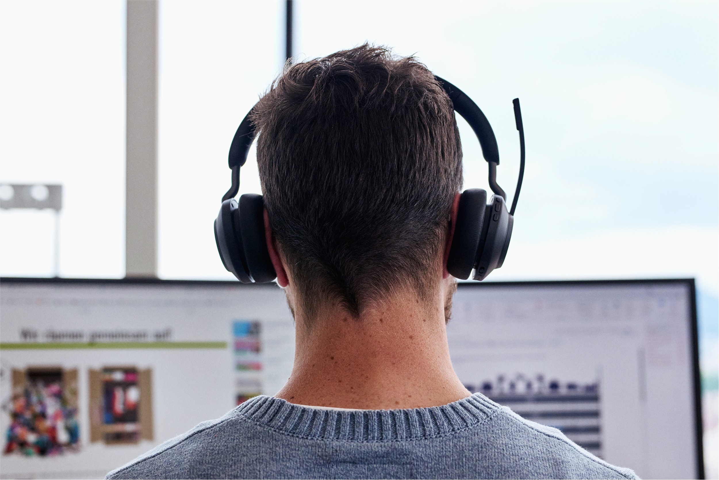 Man wearing a headset, viewed from behind, working on dual monitors in a bright office.