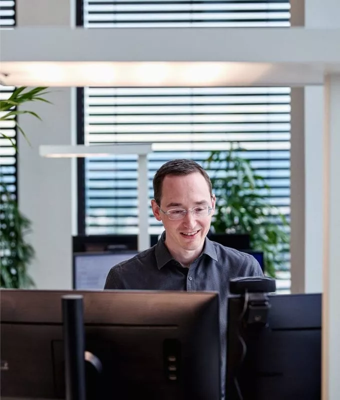 Man smiling while working at a computer in a modern office with large windows and plants.