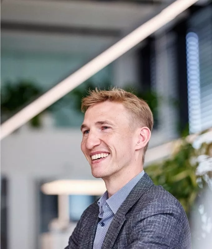 Smiling man in a grey blazer and blue shirt in a modern office setting with plants and natural light.