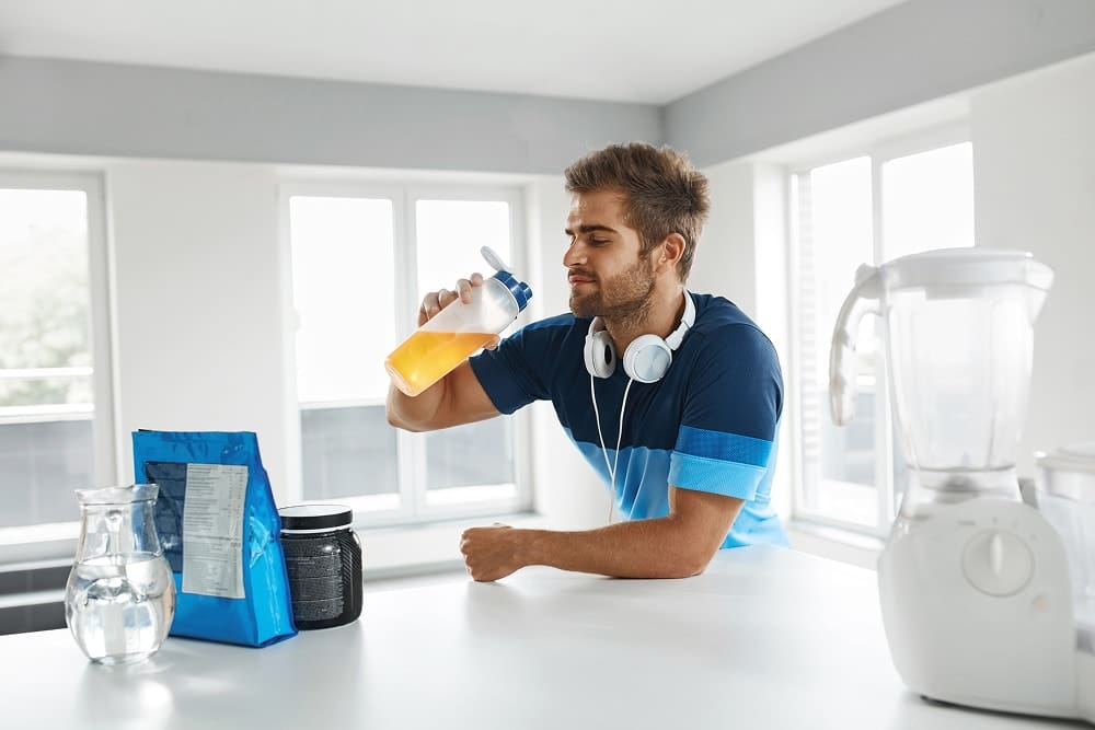 Man in sportswear holding a shaker bottle with orange drink in a bright kitchen.