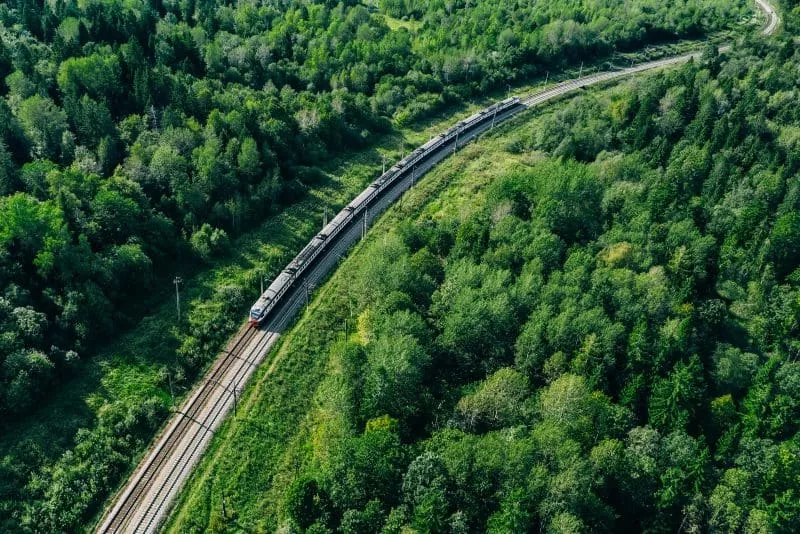 A long passenger train winds through a lush green forest, following a curved railway track.