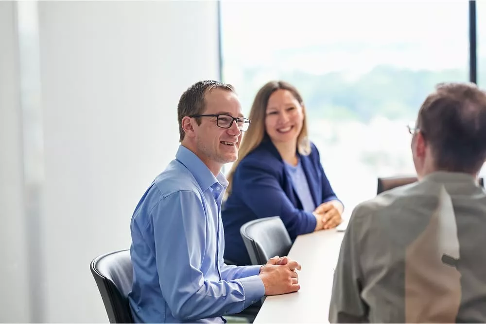 Three professionals, two men and one woman, sit around a conference table in a bright office space, smiling and engaged in conversation. image