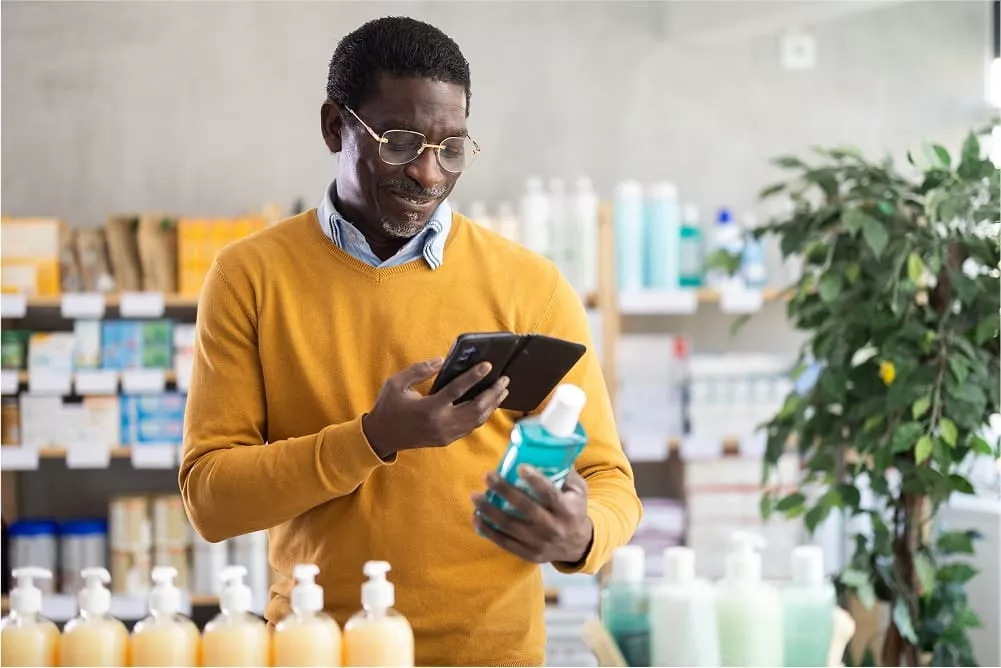 Man wearing a mustard yellow sweater reading the label of a mouthwash bottle while checking information on his smartphone in a store aisle stocked with personal care products. image