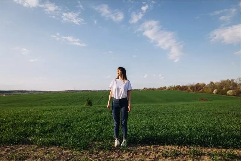 Woman standing in a green field under a blue sky with scattered clouds, wearing a white T-shirt and jeans, looking off into the distance.