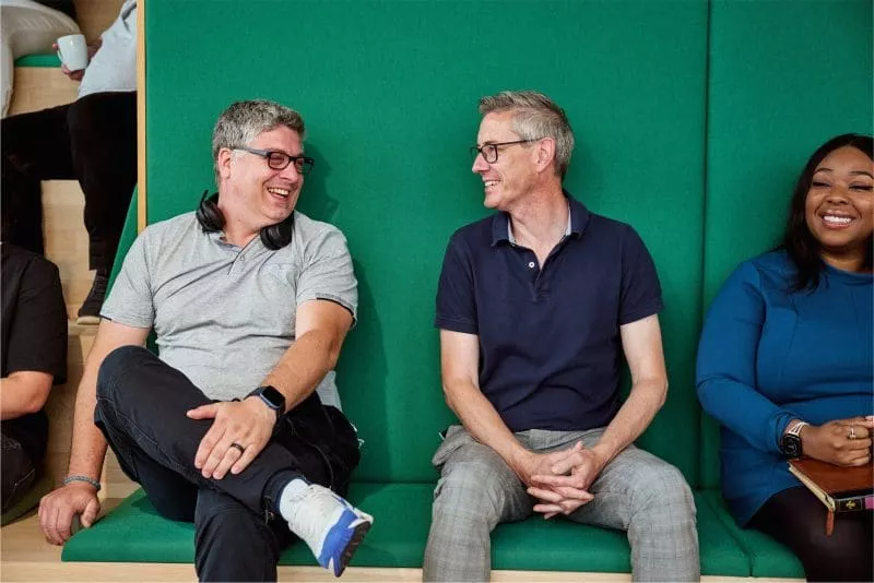 Three colleagues sitting on a green bench, smiling and chatting.