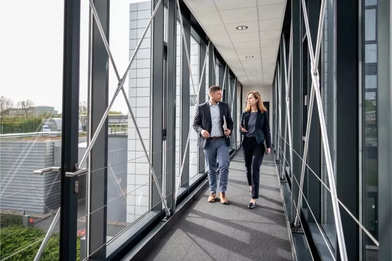 Two business professionals, a man and a woman, walk and talk in a glass-enclosed skywalk connecting modern office buildings.