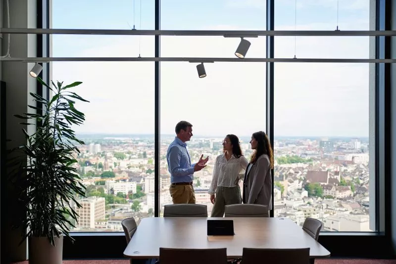 Three colleagues standing and talking in a high-rise conference room with large windows overlooking a sprawling cityscape.