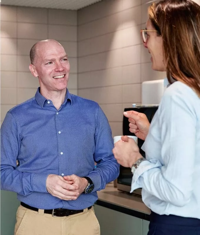 Three colleagues chatting and smiling in an office kitchen area, holding coffee cups.