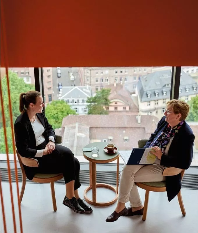 Two women having a conversation in a modern office lounge with large windows overlooking city rooftops, framed by orange vertical bars.