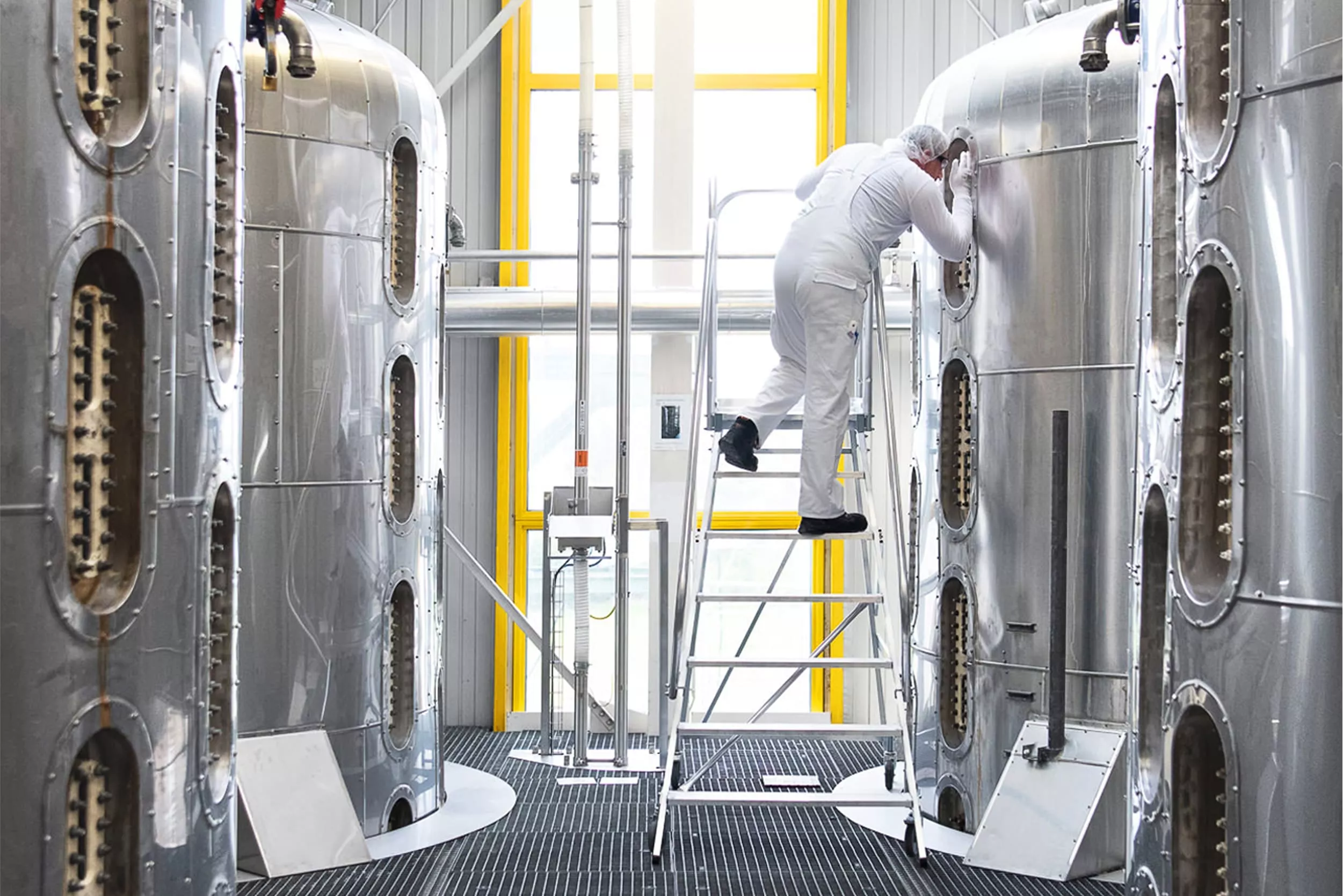 A worker dressed in white protective clothing stands on a mobile ladder, inspecting the inside of a large stainless steel industrial tank in a clean and modern processing facility. image