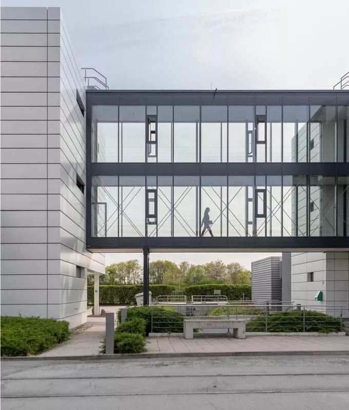 Person walking through a modern glass skywalk connecting two office buildings, with greenery visible in the background.