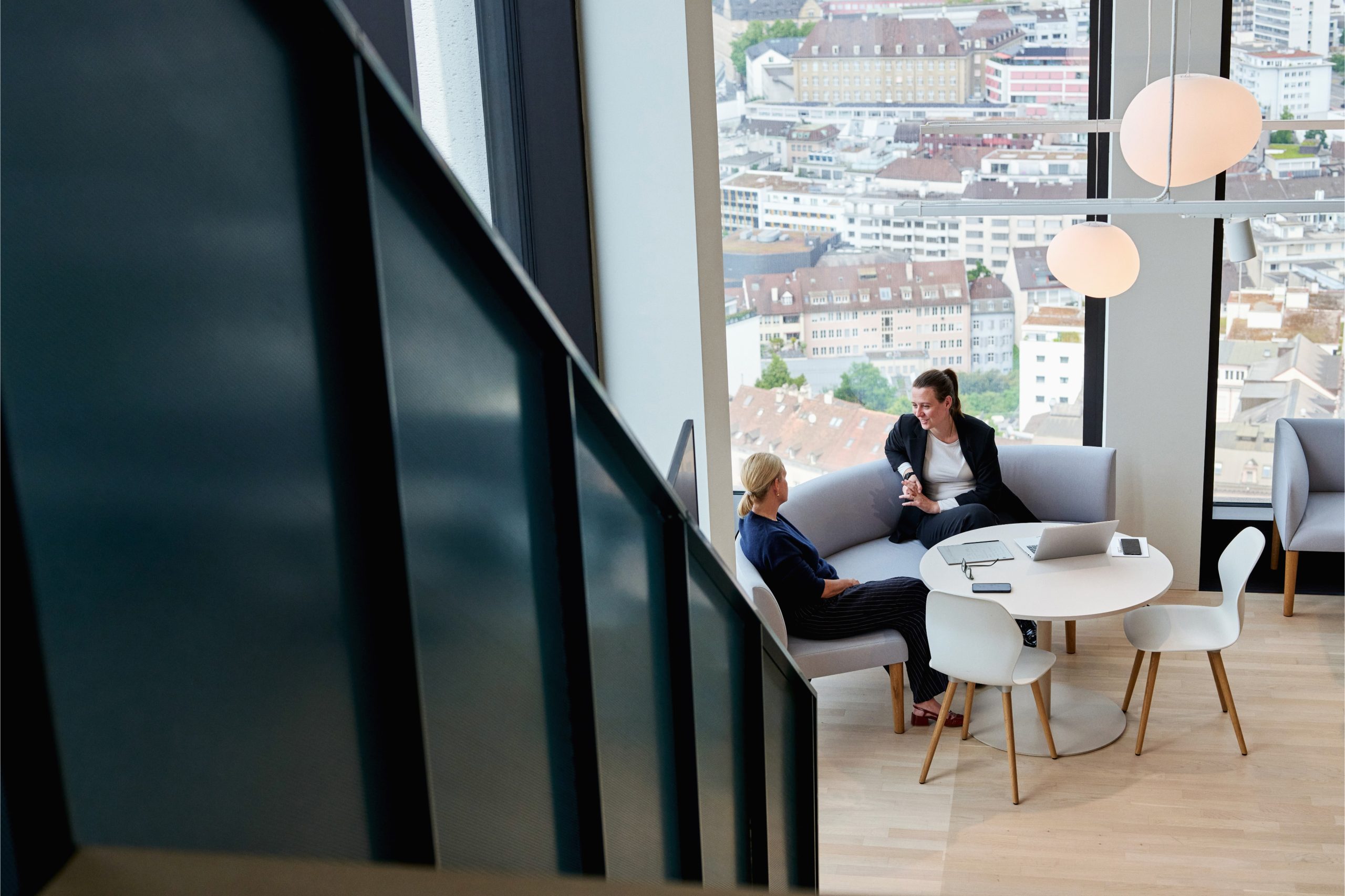 Two women in business attire having a focused discussion in a modern, high-rise office lounge with large windows offering a panoramic view of a cityscape.