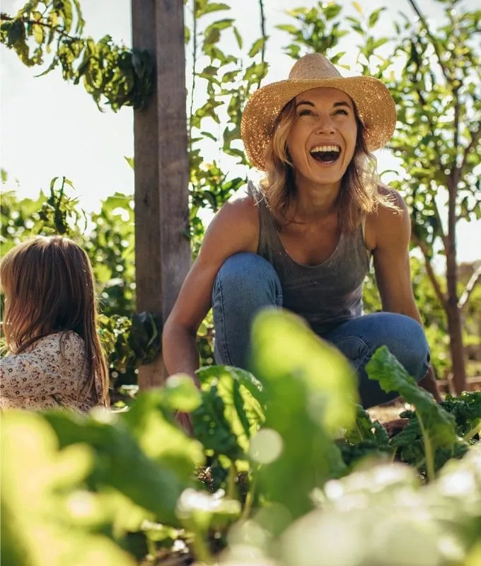 Woman in a straw hat laughing while gardening with a young girl among leafy green plants on a sunny day.