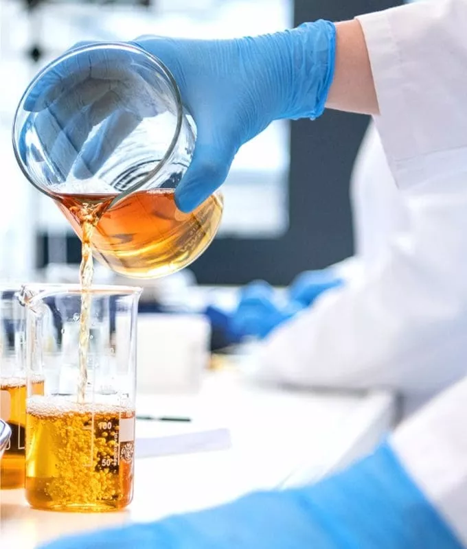 Gloved hand pouring amber liquid from one beaker to another in a laboratory.