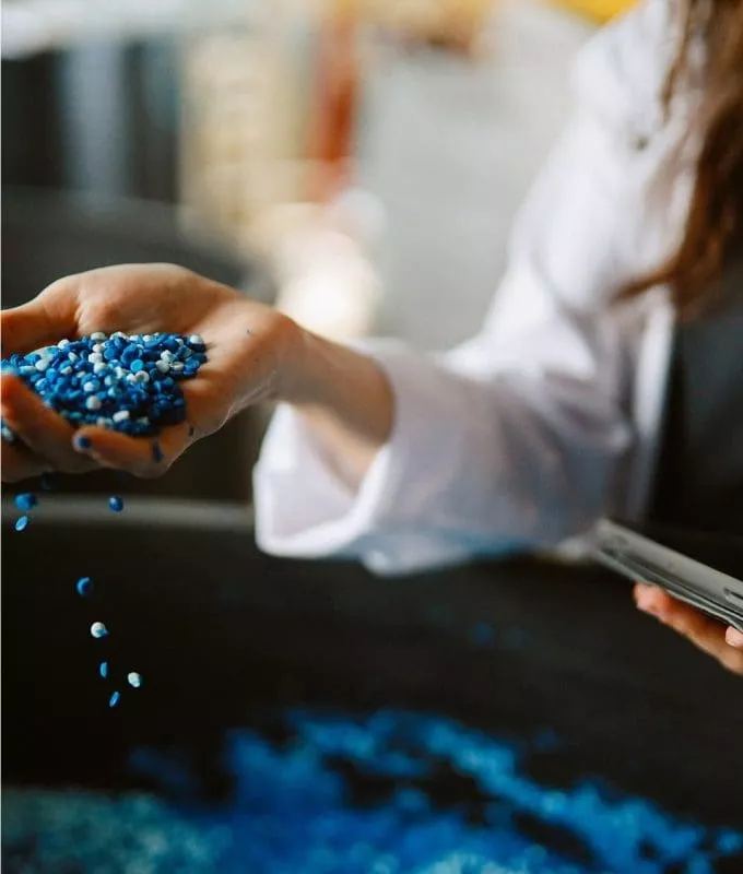 Person in lab coat holding and pouring blue plastic pellets while holding a tablet.