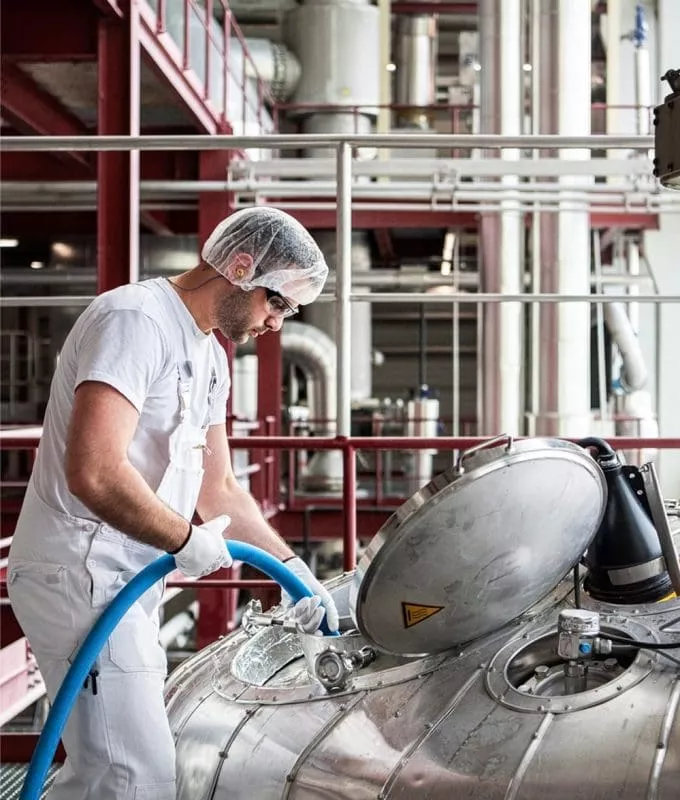 Worker in protective gear handling a hose connected to industrial stainless steel equipment in a factory.