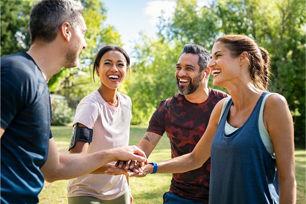 Group of four adults in sportswear laughing and stacking hands outdoors in a sunny park